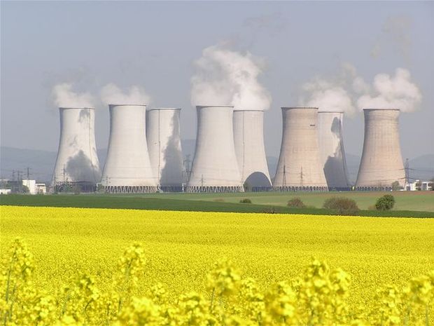 Niz velikih rashladnih tornjeva iz elektrane ispušta paru u zrak. U prvom planu, ogromno polje jarkog žutog cvijeća u kontrastu je s industrijskim strukturama u pozadini. Nebo je vedro, što pridonosi mirnoj, ali industrijskoj atmosferi slike.