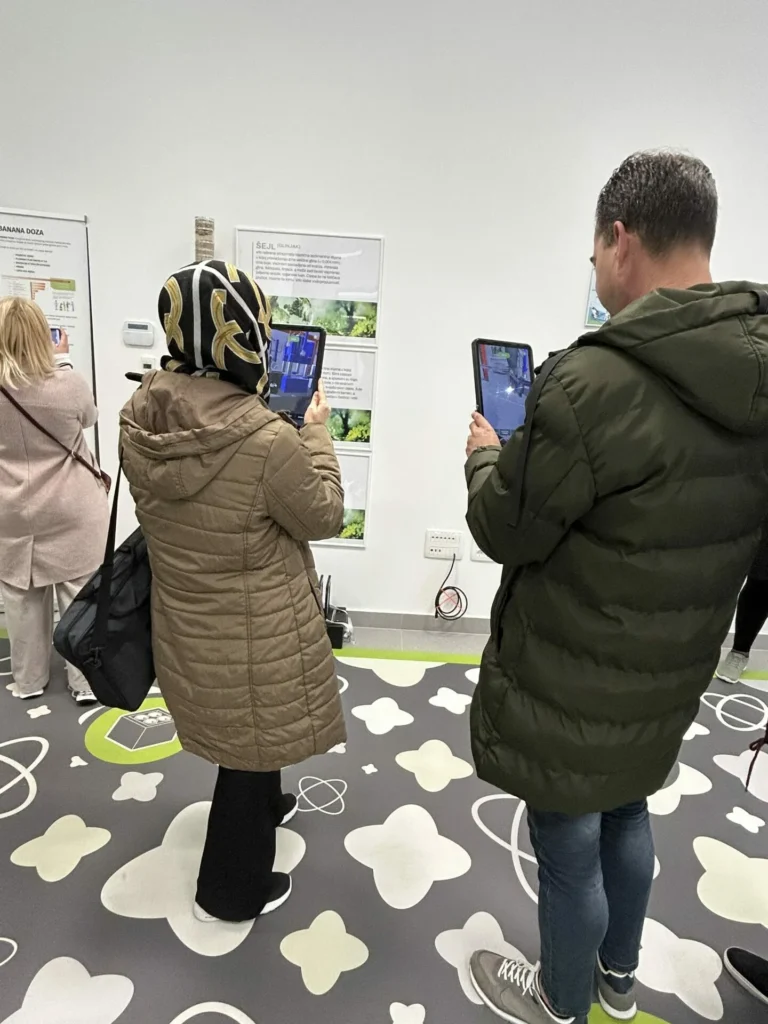 Dvije osobe stoje na podu s uzorkom, svaka drži tablet i gleda obrazovni zaslon na zidu. Jedna nosi maramu s uzorkom i smeđu jaknu, a druga nosi zelenu jaknu. Više ljudi je u pozadini.