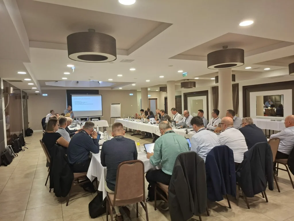 Grupa ljudi sjedi oko velikog konferencijskog stola u obliku slova U u sobi za sastanke i gleda prezentaciju na ekranu sprijeda. Nekoliko njih ima otvorena prijenosna računala i uključeno je u raspravu.