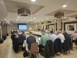 Grupa ljudi sjedi oko velikog konferencijskog stola u obliku slova U u sobi za sastanke i gleda prezentaciju na ekranu sprijeda. Nekoliko njih ima otvorena prijenosna računala i uključeno je u raspravu.
