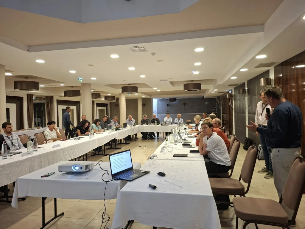 Grupa ljudi sjedi oko velikog stola u obliku slova U u konferencijskoj dvorani i sluša govornika koji stoji s desne strane. Prijenosno računalo i projektor se uvlače naprijed.