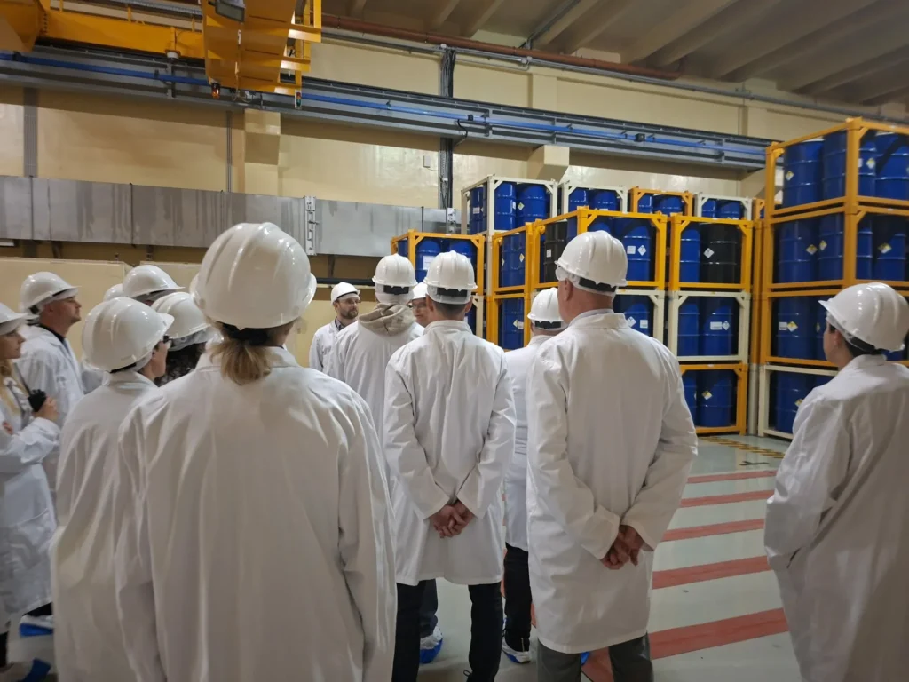 Skupina ljudi u bijelim laboratorijskim kutama i zaštitnim kacigama stoji u skladištu, okrenuta prema naslaganim plavim bačvama na žutim policama, slušajući osobu koja kao da održava obilazak ili prezentaciju.