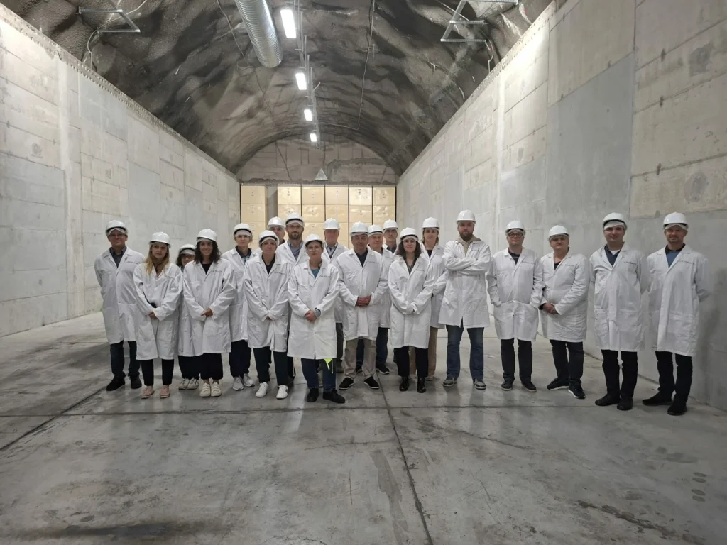 Grupa ljudi u bijelim laboratorijskim kutama, zaštitnim kacigama i zaštitnim navlakama za cipele stoji zajedno u velikom, industrijskom podzemnom objektu s betonskim zidovima i stropom.