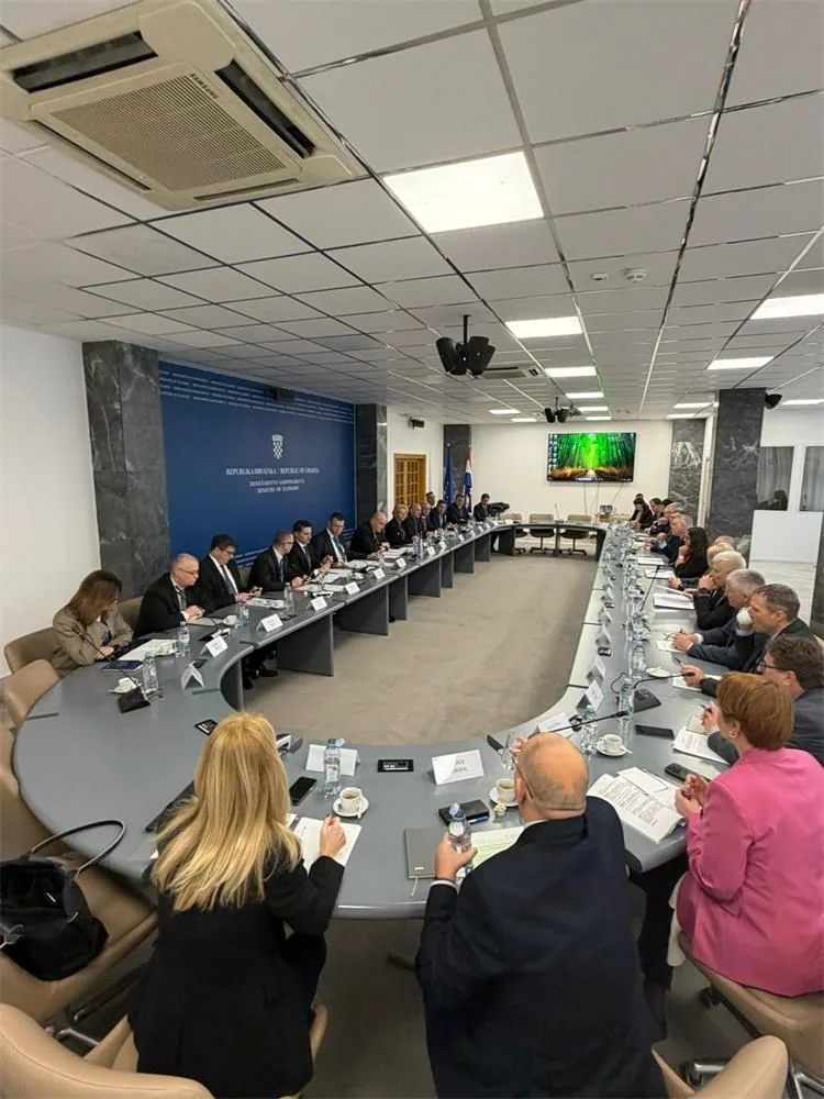 Velika skupina ljudi u poslovnoj odjeći sjedi oko dugog, ovalnog konferencijskog stola u modernoj sobi za sastanke, raspravlja o dokumentima i bilježi. Vidljivi su plavi zid s logotipom i monitor sa zelenom slikom.