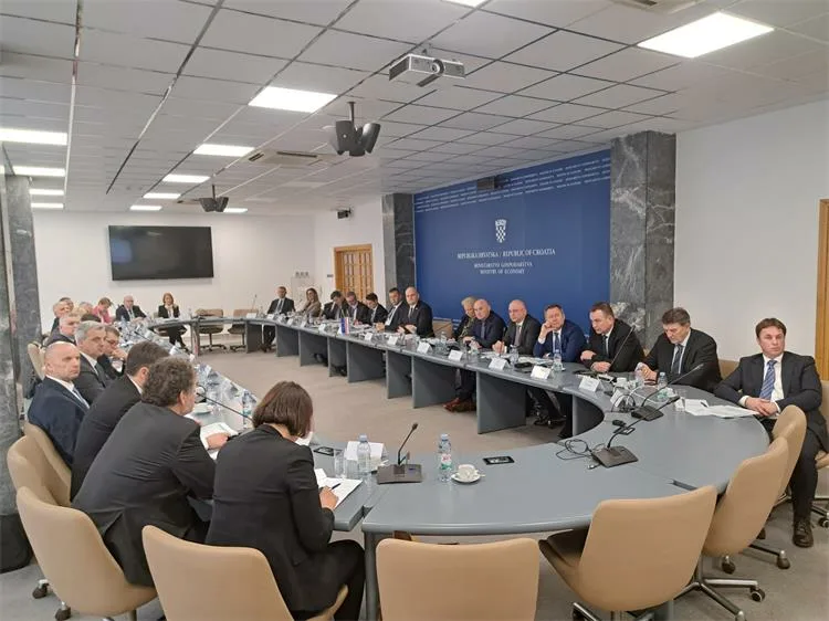 Grupa ljudi u svečanoj odjeći sjedi oko velikog konferencijskog stola u obliku slova U u sobi za sastanke s plavim zidom, mikrofonima, dokumentima i bocama vode na stolu.