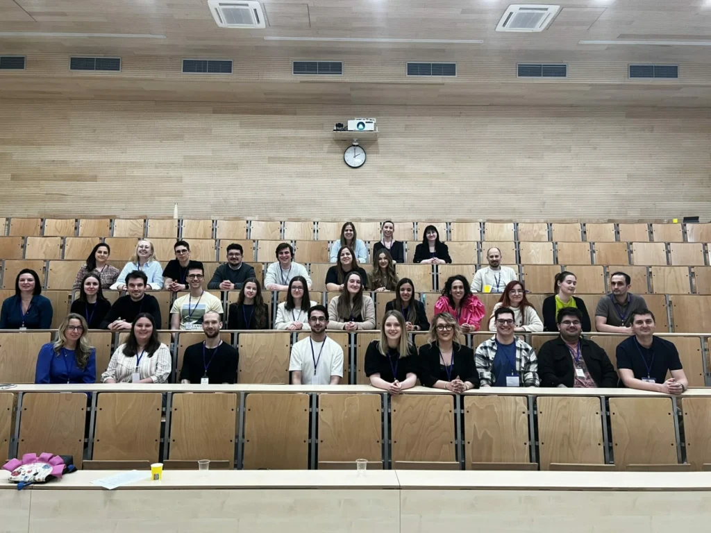 Grupa od oko 35 ljudi sjedi i stoji u praznoj predavaonici s drvenim sjedalima, pozirajući za fotografiju. Većina se smiješi i okrenuta je prema kameri; neki nose vrpce. Na stražnjem zidu vidljivi su sat i projektor.