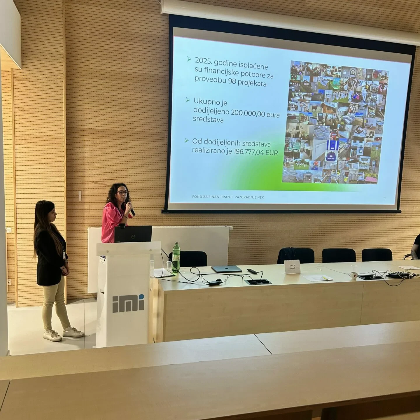 Dvije žene stoje na prednjem dijelu predavaonice i drže prezentaciju. Iza njih se projicira slajd koji prikazuje tekst i kolaž fotografija. Na stolu je postavljeno nekoliko prijenosnih računala i mikrofona.