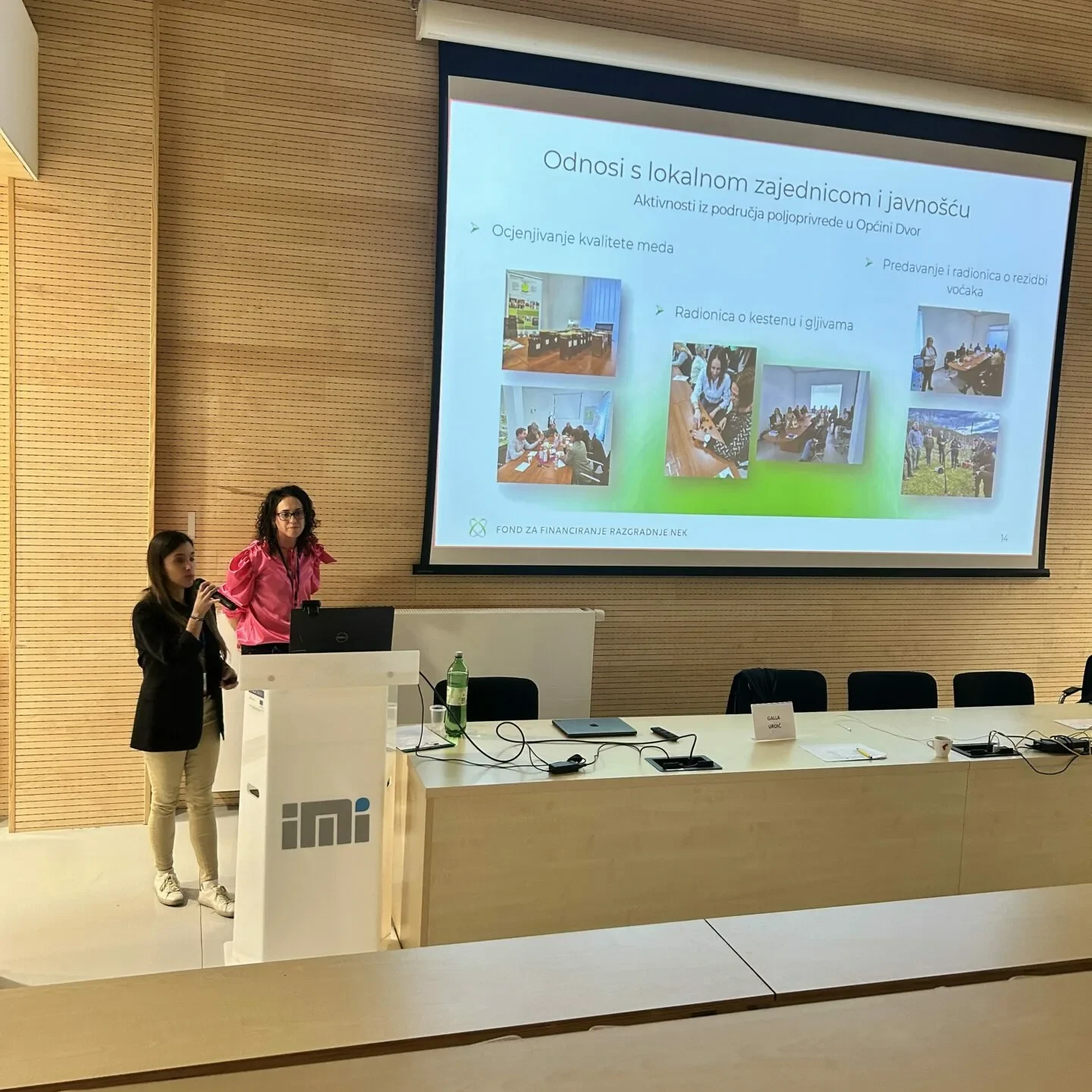 Dvije žene stoje za podijem i prezentiraju slajdove u predavaonici. Slajd na ekranu prikazuje fotografije i tekst na hrvatskom jeziku, a prostorija ima drvene zidove i moderan, minimalistički dizajn.