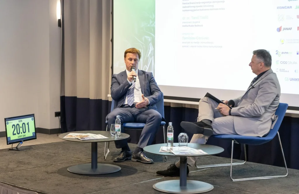 Dva muškarca u odijelima sjede na pozornici u foteljama i raspravljaju uz mikrofone. Na stolovima između njih nalaze se boce s vodom i papiri. Digitalni sat pokazuje 20:01, a iza njih se vidi prezentacijski ekran.