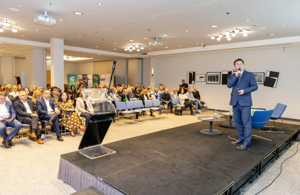 Muškarac u plavom odijelu govori na pozornici s mikrofonom publici koja sjedi u redovima na konferenciji ili seminaru. Vidljivi su podij i platno, a na zidu iza njega visi nekoliko uokvirenih slika.