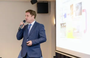 Muškarac u plavom odijelu govori u mikrofon dok drži daljinski upravljač, stojeći pored projiciranog prezentacijskog platna s dijagramima i slikama.