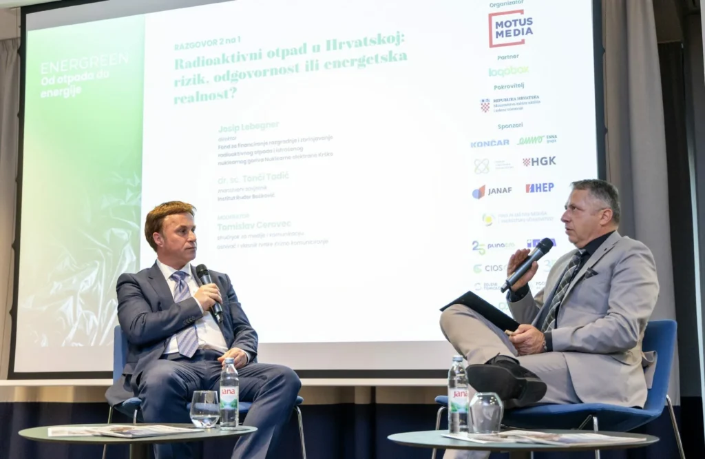 Dva muškarca u odijelima sjede na pozornici s mikrofonima i raspravljaju. Iza njih je veliki ekran na kojem se prikazuje prezentacija na hrvatskom jeziku o radioaktivnom otpadu u Hrvatskoj, s logotipima sponzora i detaljima događaja.