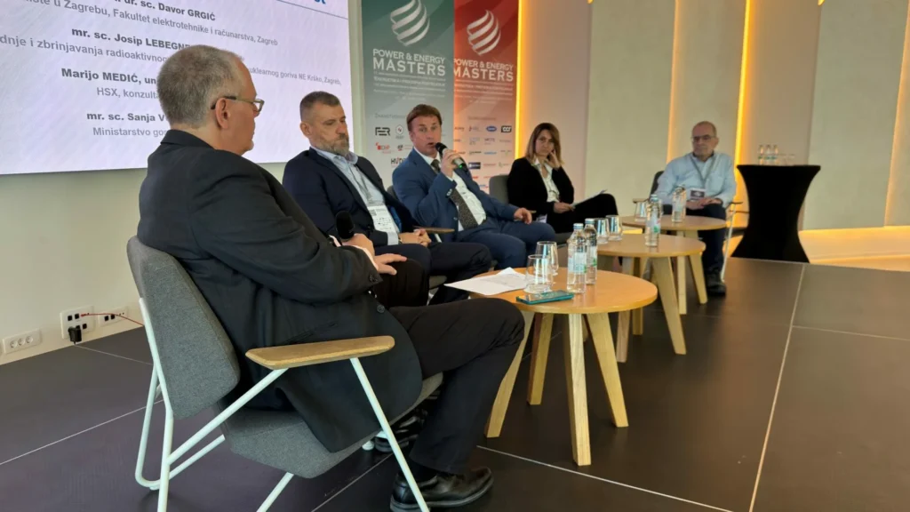 Petero ljudi sjedi na pozornici u panel raspravi na konferenciji. Četiri muškarca i jedna žena okrenuti su prema publici, s mikrofonima i čašama za vodu na malim stolovima između njih. U pozadini je vidljiv transparent "Power & Energy Masters".
