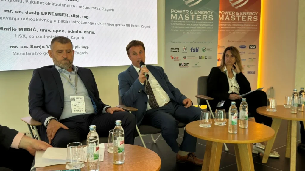 Tri panelista u poslovnoj odjeći sjede na pozornici tijekom događaja Power & Energy Masters, s mikrofonima i bocama vode na stolu. Iza njih vidljivi su veliki prezentacijski ekran i transparenti događaja.