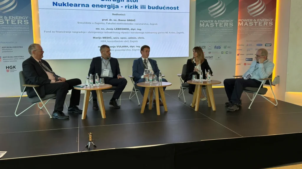 Petero ljudi sjedi na pozornici tijekom panel rasprave na konferenciji, s ekranom iza njih koji prikazuje informacije o nuklearnoj energiji. Mali stolovi s bocama vode nalaze se ispred govornika.